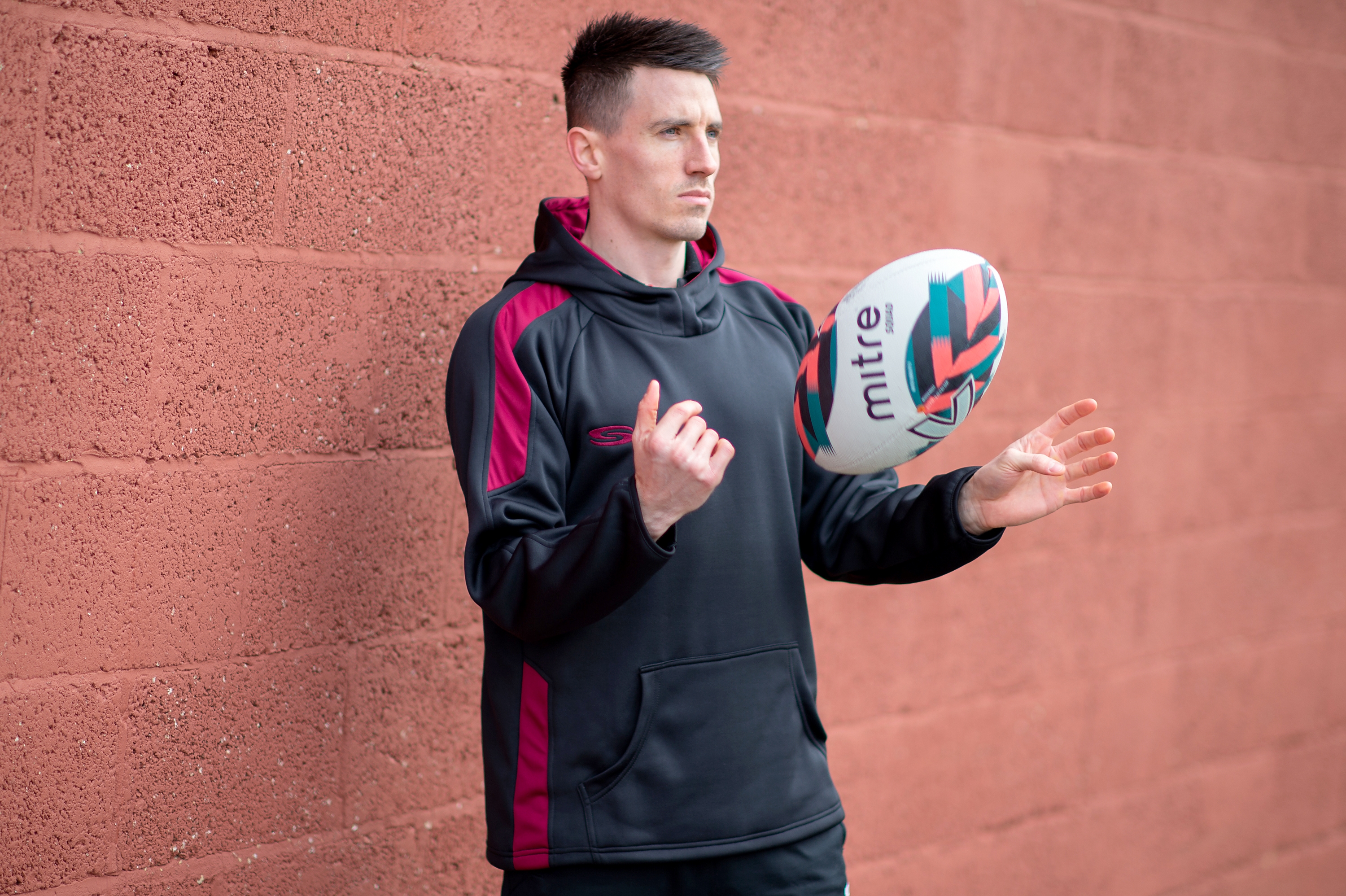 Teaser image for our teamwear solution linker Male in black/maroon hoodie spinning a rugby ball in the air